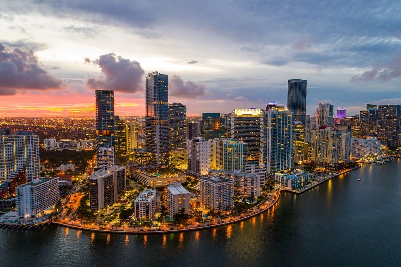 A vibrant cityscape at dusk, featuring tall, illuminated skyscrapers lining a waterfront. The sky is painted with shades of orange, pink, and blue as the sun sets in the background. The water below reflects the buildings and the sky above.